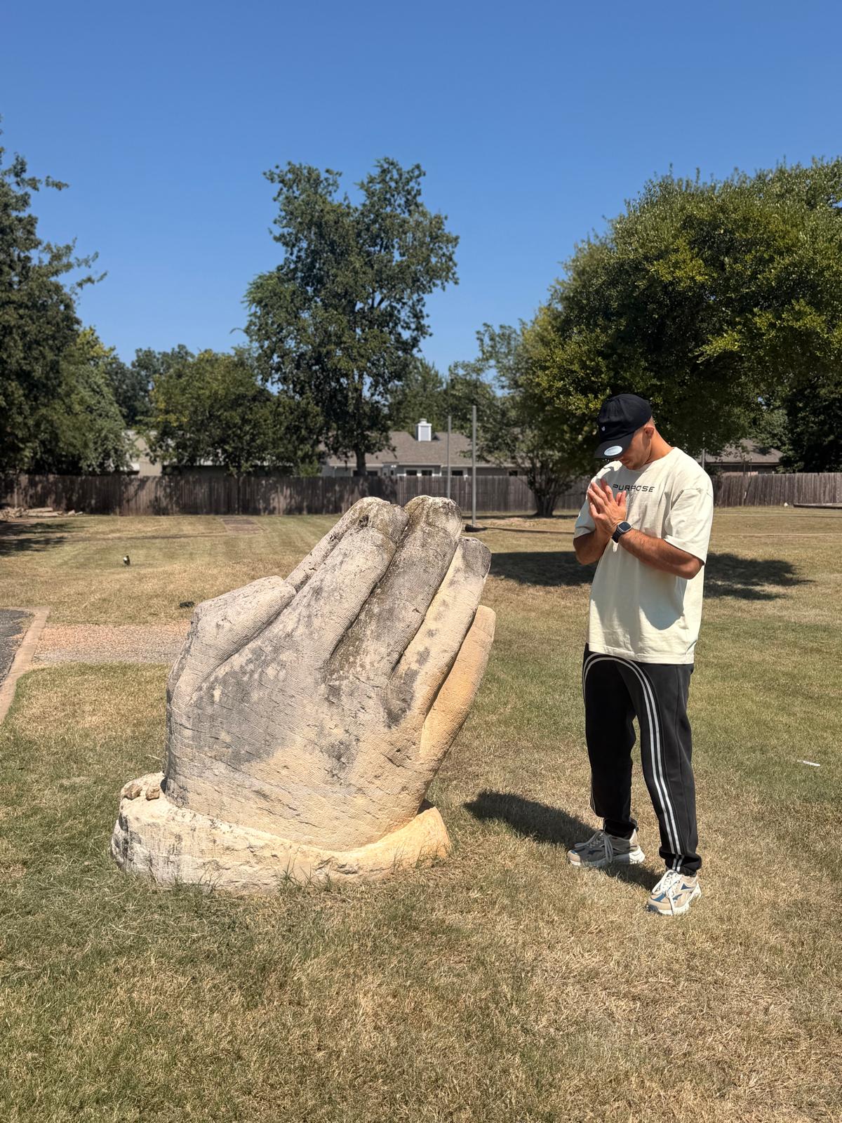Hands together in front of a stone praying-hands sculpture, wearing the Purpose Tee